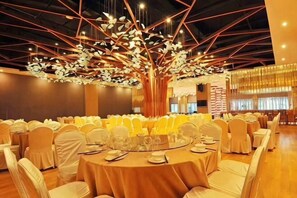 Una sala per banchetti con tavoli rotondi, sedie bianche e un grande albero artificiale come elemento decorativo.