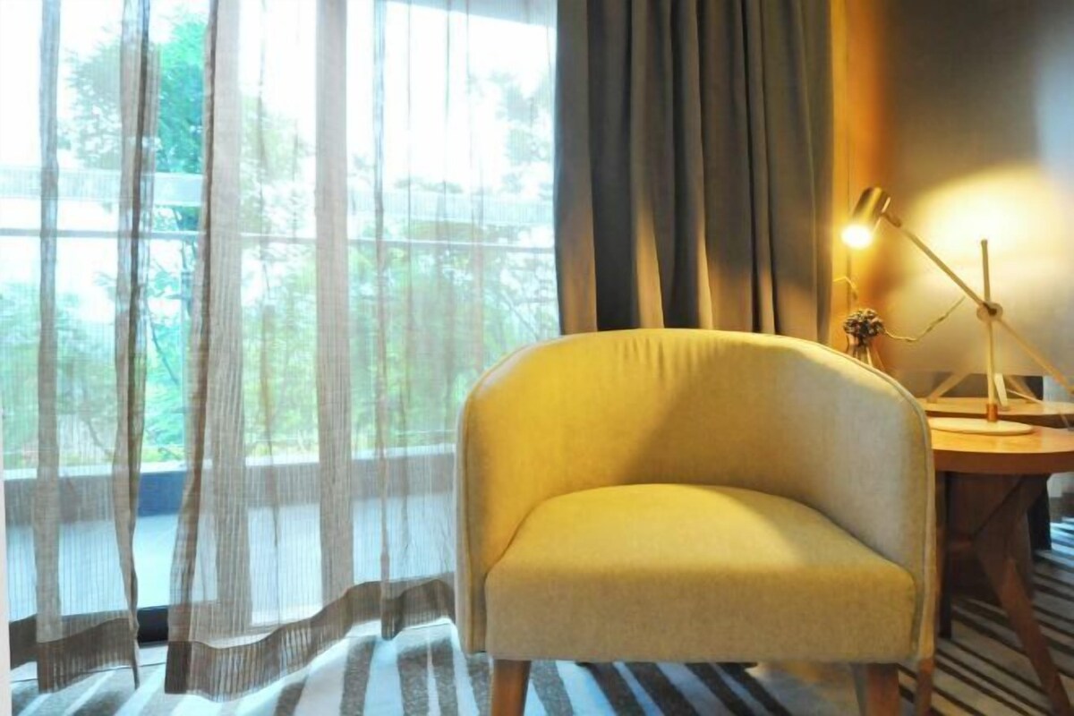 Una habitación con un sillón amarillo, un escritorio con lámpara y cortinas translúcidas que dejan entrar luz natural.