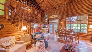 A spacious log cabin interior with a dining area, a living room with a sofa and armchair, a kitchen, and a staircase leading to an upper level.