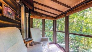 A cabin with two upholstered chairs, a wooden table, and a view of the forest through large windows.