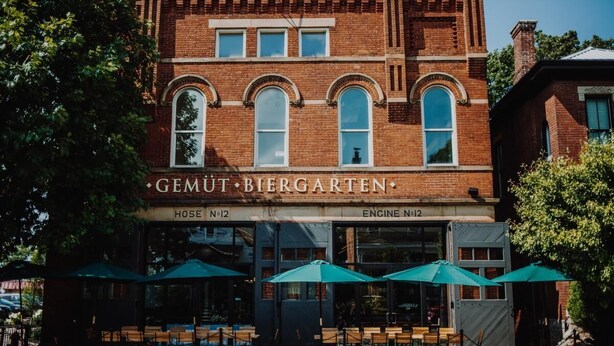 A brick building with the name "GEMUT BIERGARTEN" and "HOSE No.12" and "ENGINE No.12" signs. There are outdoor tables with green umbrellas.