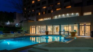 Una piscina con escalones que bajan hasta la orilla, un edificio de varios pisos y ventanas, y un área de entrada bien iluminada.
