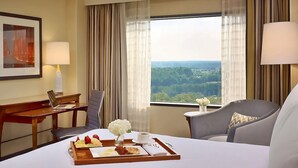 Habitación de hotel con cama, escritorio, silla, mesa de comedor con desayuno y un ventanal con vistas.