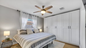 Ein Schlafzimmer mit einem Bett, einem Deckenventilator, weißen Schränken und einem Fenster mit durchsichtigen Vorhängen.