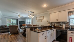Une cuisine moderne avec des armoires blanches, un comptoir noir et un lave-vaisselle encastré. On y trouve un coin repas avec une table et des chaises, et un ventilateur de plafond au-dessus.
