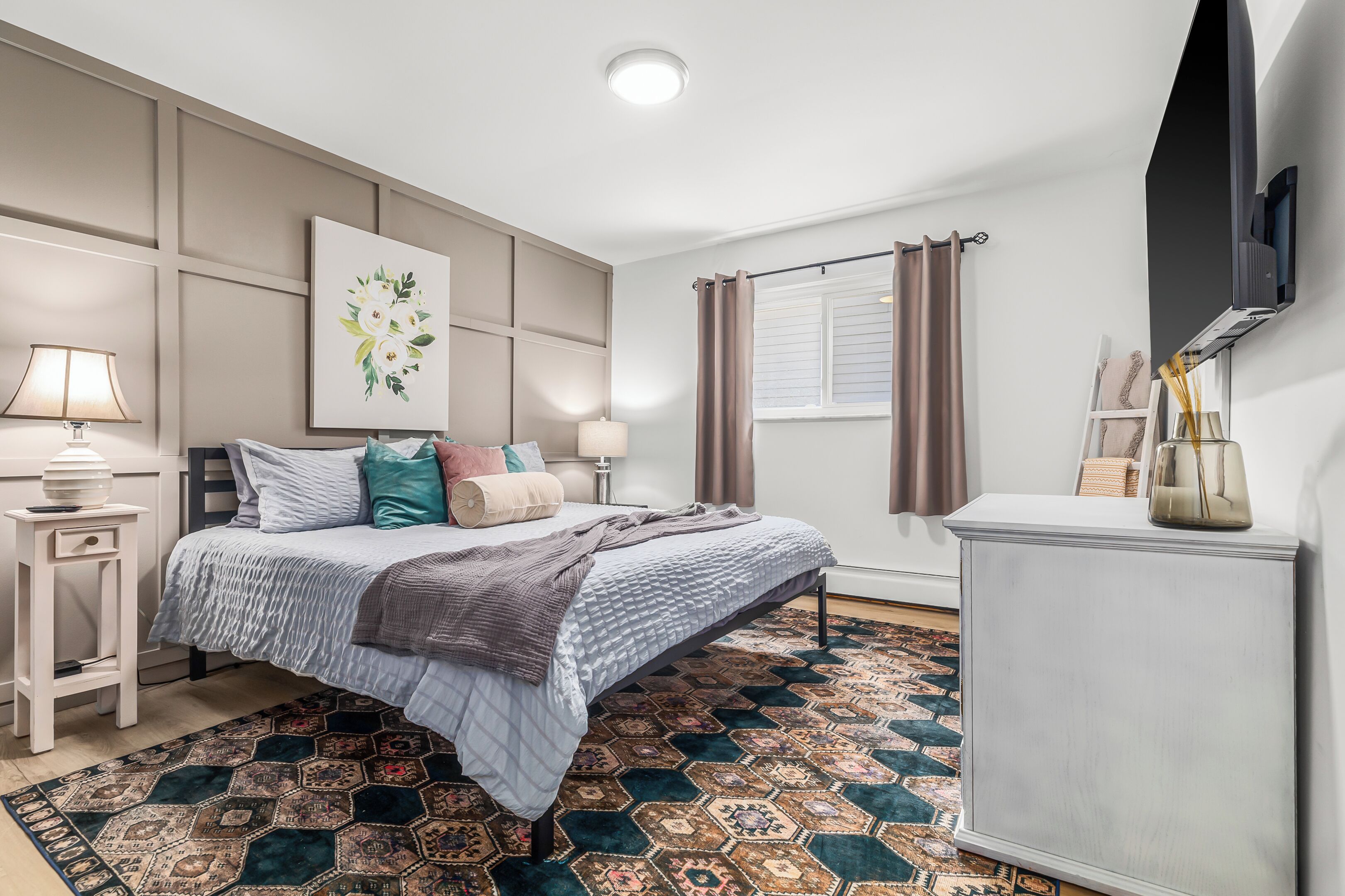 A bedroom with a bed, bedside tables, a rug, a painting, a TV, and a window with curtains.