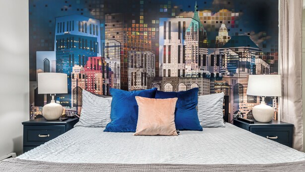 A bed with a cityscape mural on the wall behind it, two bedside tables with lamps, and a textured bedspread. - Bright & Spacious 2 Bedroom | Quiet | Safe | Clean (Mount Clemens)
