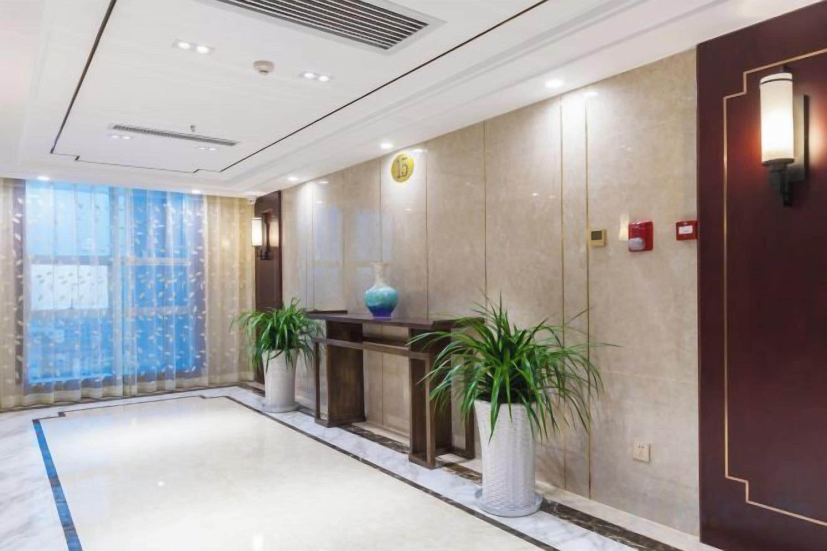 A hotel lobby with a reception desk, potted plants, a large vase, and a red door.
