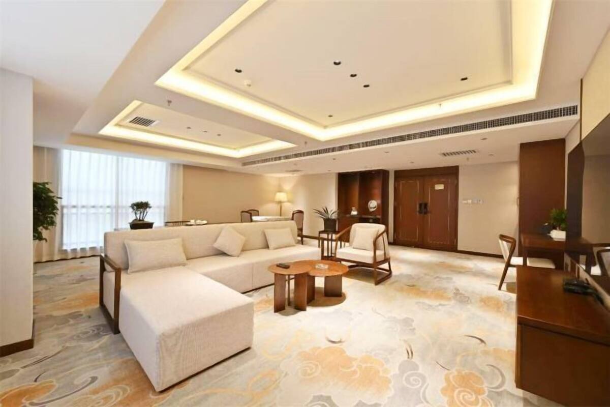 A modern hotel room with a sectional sofa, a round coffee table, and a dining area with a table and chairs.