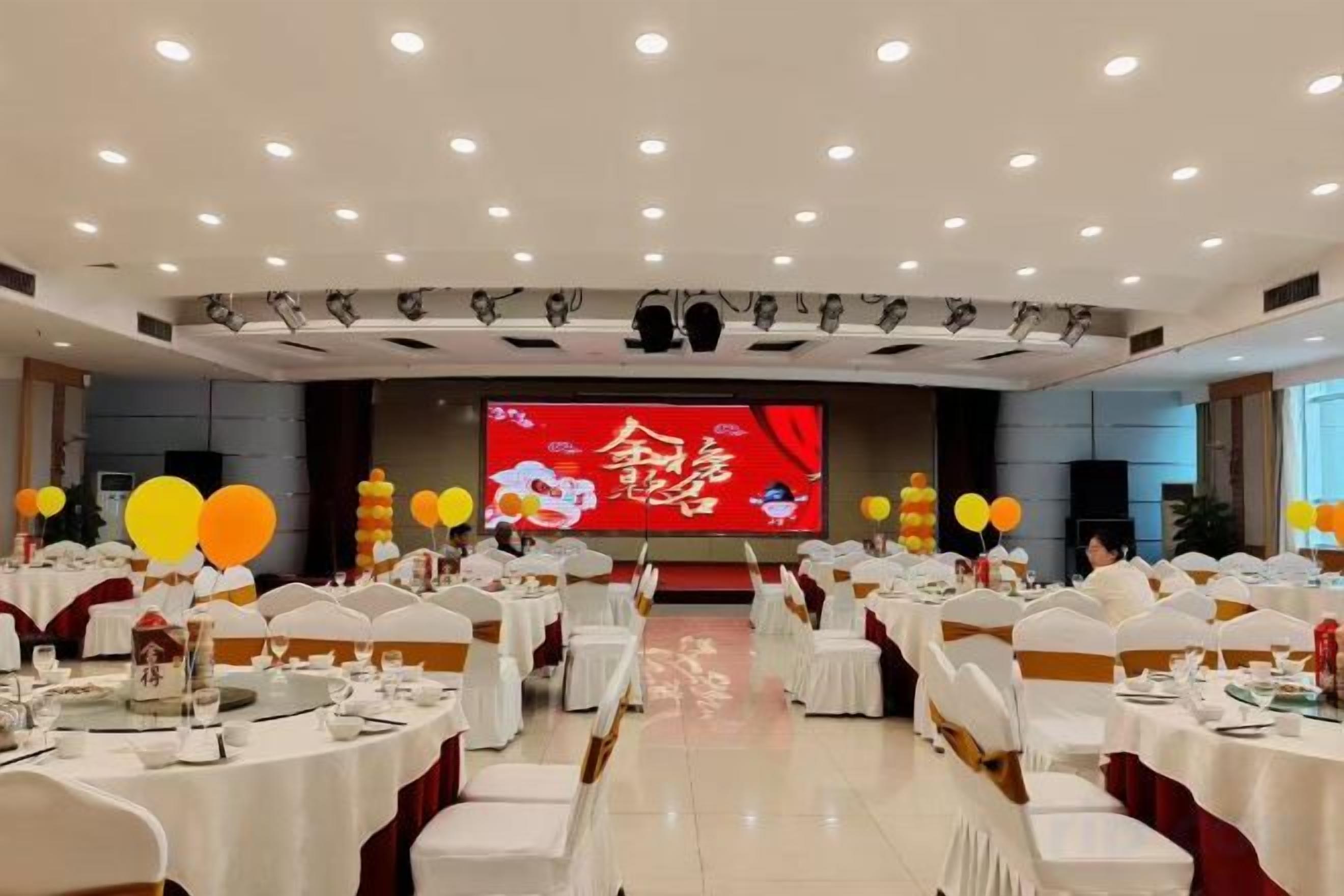 Un salón de banquetes con mesas redondas, sillas blancas y un escenario con una pantalla grande.