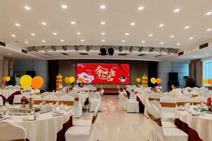 Un salón de banquetes con mesas redondas, sillas blancas y un escenario con una pantalla grande.