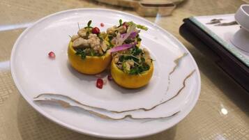 A white plate with two stuffed yellow peppers garnished with nuts and vegetables, placed on a table with a patterned tablecloth.