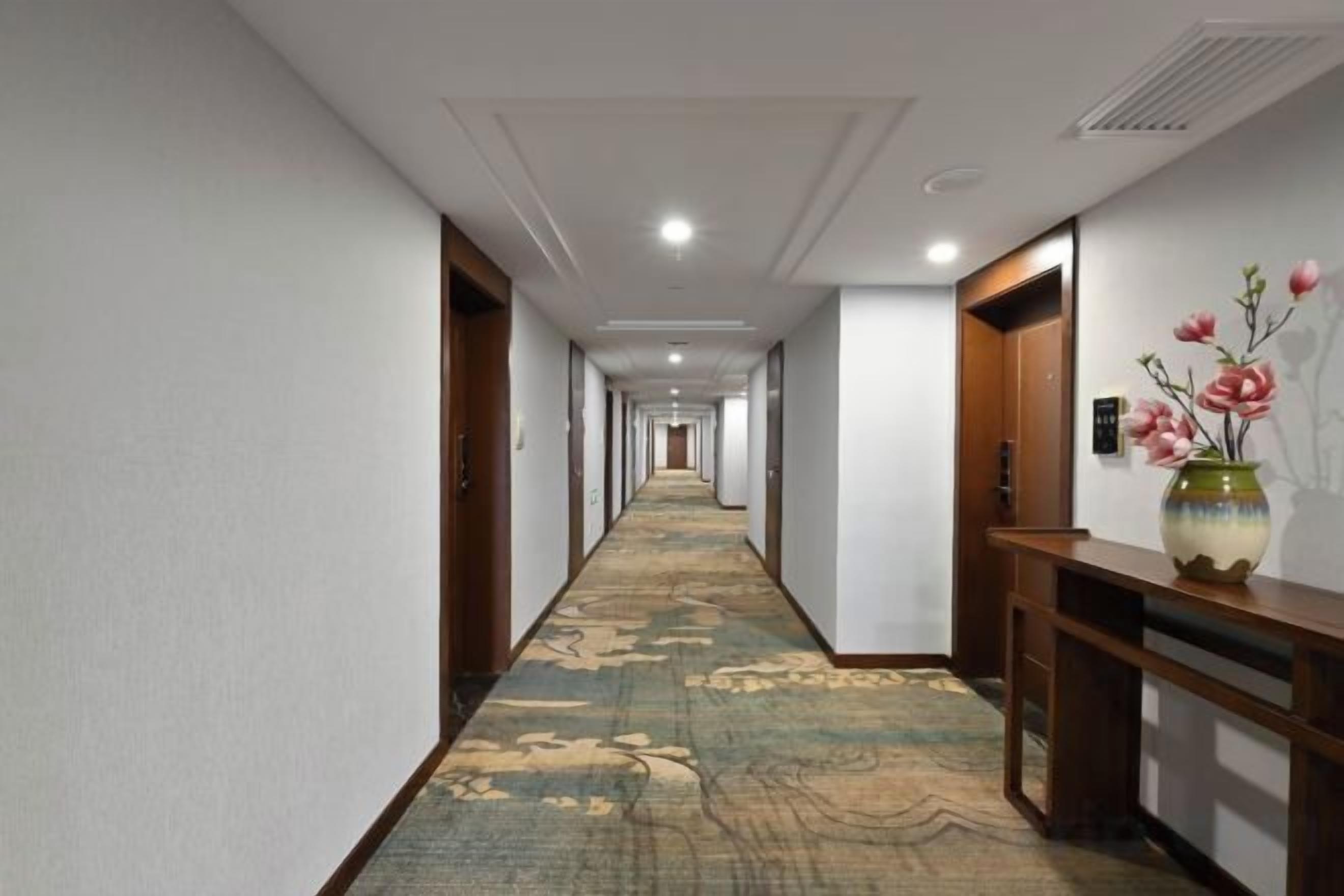Un pasillo de hotel con piso alfombrado, puertas de madera y un florero sobre una consola.