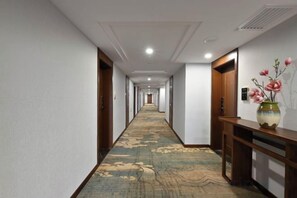 Un couloir d'hôtel avec un sol recouvert de moquette, des portes en bois et un vase rempli de fleurs posé sur une console.