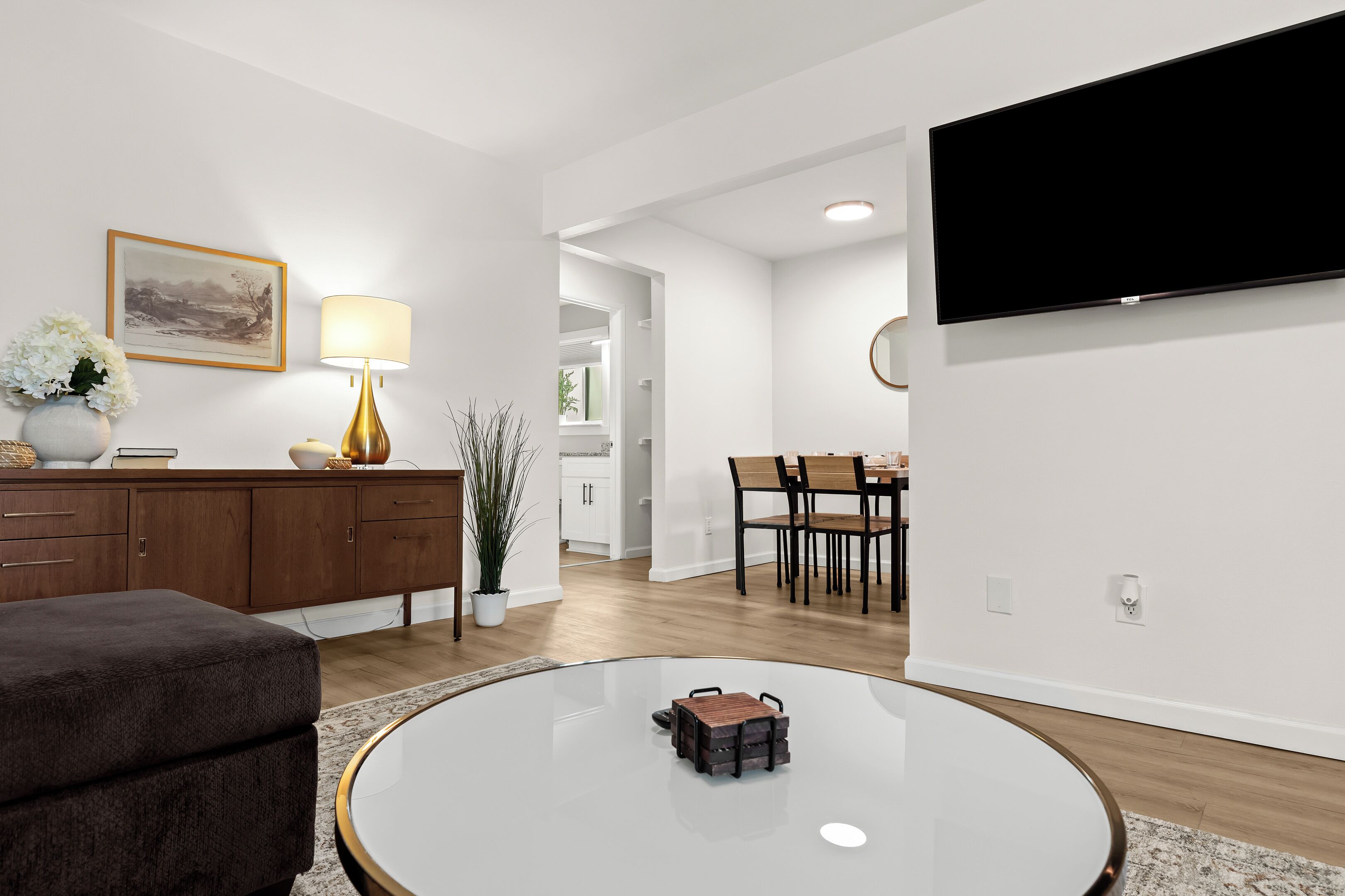 Een moderne woonkamer met een ronde glazen salontafel, een bank, een eethoek met stoelen, een flatscreen televisie aan de muur en een houten kast met een lamp.