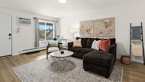 Een moderne woonkamer met een bruine bank, een witte salontafel en een vloerkleed met een patroon. Er is een raam met uitzicht op een huis, een lamp en een schilderij aan de muur.