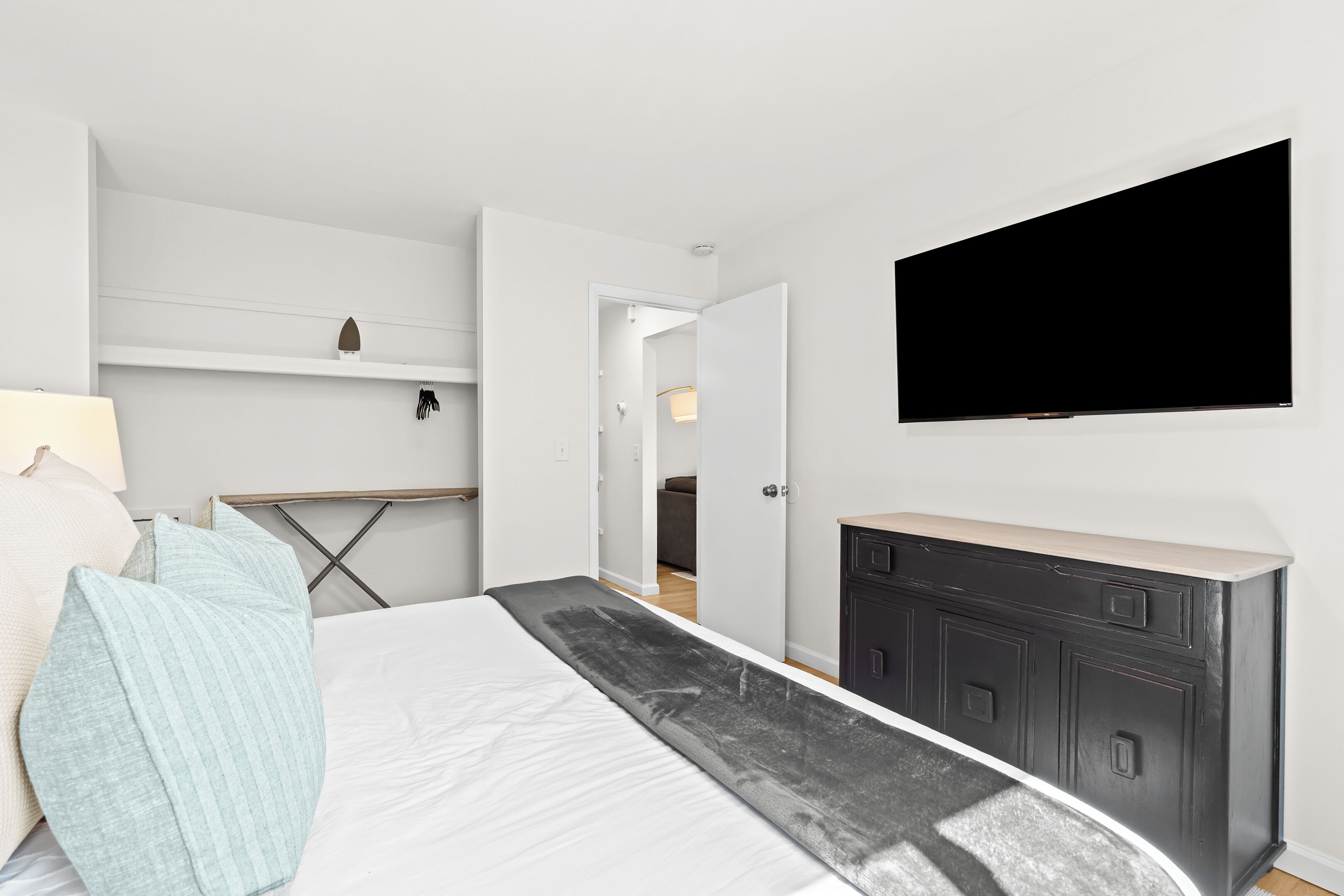 A modern bedroom with a bed, a flat-screen TV mounted on the wall, a dresser, and a desk with a lamp.