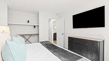 A modern bedroom with a bed, a flat-screen TV mounted on the wall, a dresser, and a desk with a lamp.