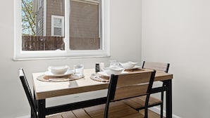 A dining table set with white bowls, glasses, and cutlery, positioned near a window with a view of a house and a wooden fence. - Contemporary 1BR, Fast WiFi, Pet-Friendly (Mount Clemens)