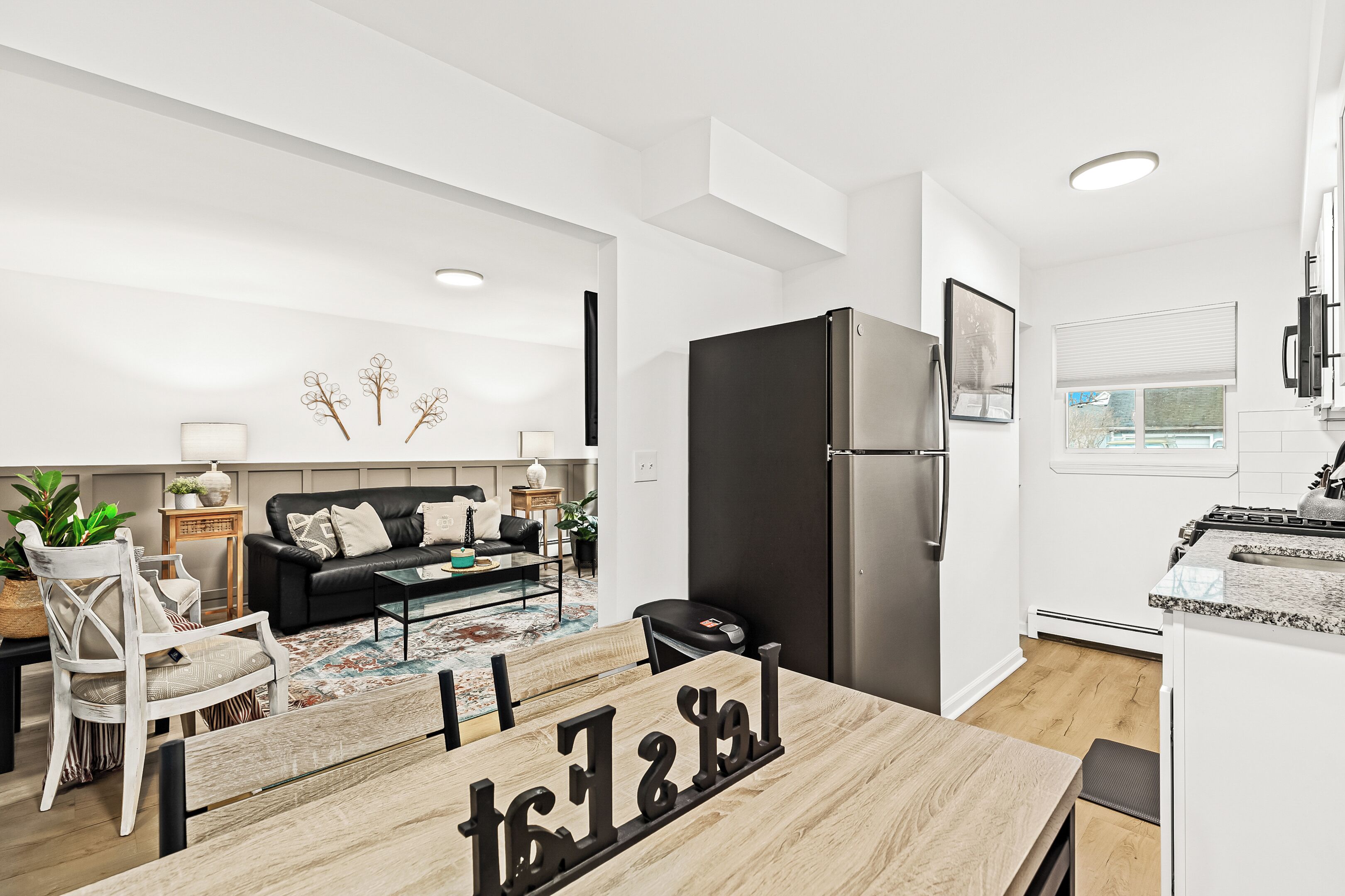 A modern kitchen with a black refrigerator, a dining area with a wooden table, and a living room with a black sofa.