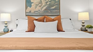 Una cama bien hecha con almohadas blancas y marrones, una manta beige y un cuadro de nubes.