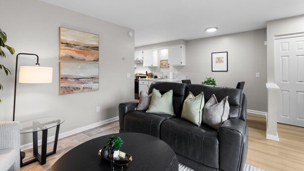 A modern living room with a black leather sofa, a glass coffee table, and a dining table. A kitchen is visible in the background with white cabinets and a countertop.