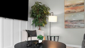 A modern living room with a round black table, a potted plant, a floor lamp, and a flat-screen TV mounted on the wall.