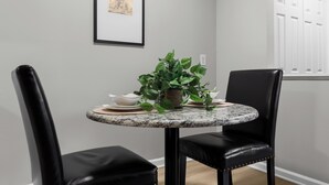 A dining area with a round table, two black chairs, a potted plant, and a framed picture on the wall.