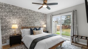 A bedroom with a large bed, two bedside tables with lamps, a ceiling fan, a view of the outdoors through a sliding glass door, and a patterned rug.
