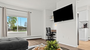 Een moderne woonkamer met een grijze bank, een glazen salontafel met een plant, een flatscreen televisie aan de muur en een uitzicht op het groen door grote ramen.