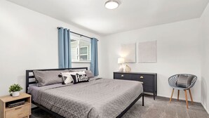 Un dormitorio moderno con cama, mesitas de noche, una silla y un ventanal con cortinas.