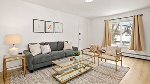 Una sala de estar moderna con un sofá gris, una mesa de centro de vidrio, un sillón color beige y un tapete estampado. Se observan obras de arte enmarcadas en la pared y una ventana con cortinas.