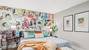 Un dormitorio con cama, una mesita de noche, una pared decorada con ilustraciones tipo cómic y dos cuadros enmarcados.
