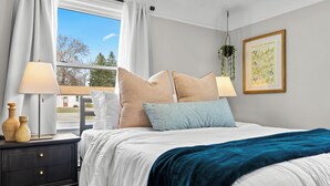 Un dormitorio con una cama grande, ropa de cama blanca, una mesita de noche oscura con dos lámparas e impresión enmarcadas de motivos botánicos en la pared.