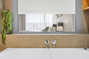 A modern bathroom with a bathtub, a window with a view of a bed and a lamp, and a green plant on the left.