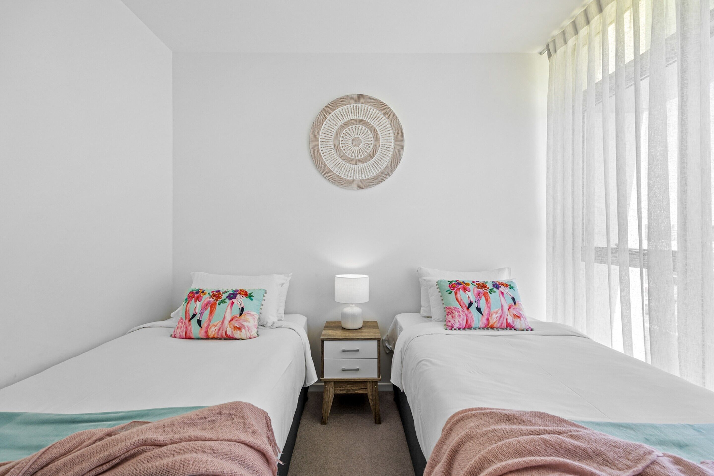 A hotel room with two beds, a bedside table with a lamp, a decorative wall hanging, and a window with curtains.