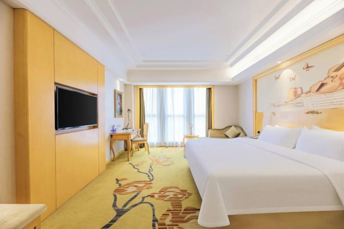 Una habitación de hotel con cama, escritorio, televisor y ventana con cortinas.