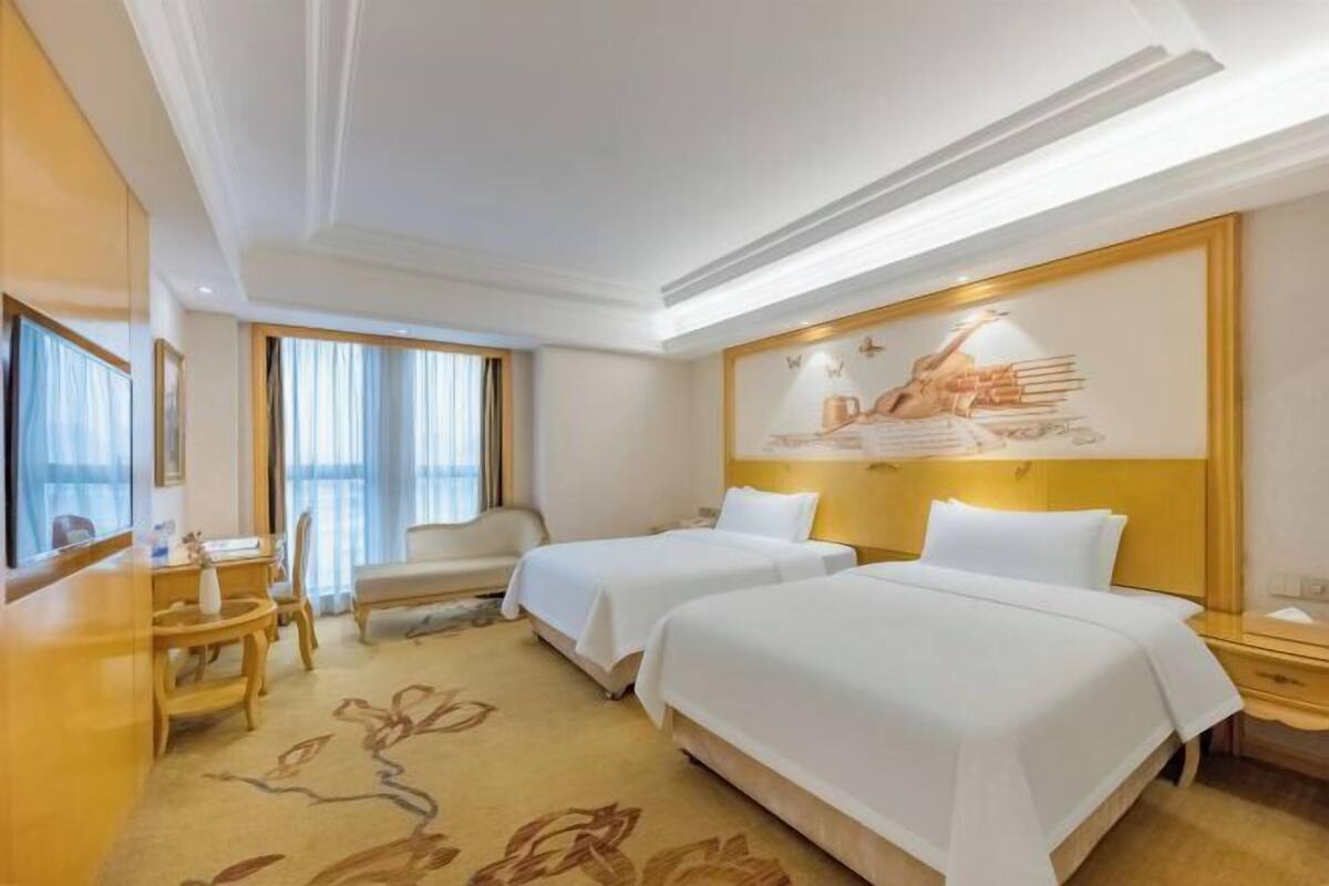 A hotel room with two beds, a desk, a chair, and a large window with curtains.