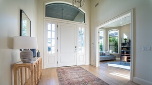 Ein geräumiger Eingangsbereich mit einem Holzkonsolentisch, einem großen Teppich und einer Doppeltür, die zu einem Wohnbereich mit Sofa und Bücherregalen führt.