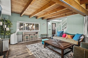 Ein gemütliches Wohnzimmer mit Holzbalkendecke, einer Couch mit bunten Kissen, einem Couchtisch und einer kleinen Küchenzeile mit Fenster und Ventilator.