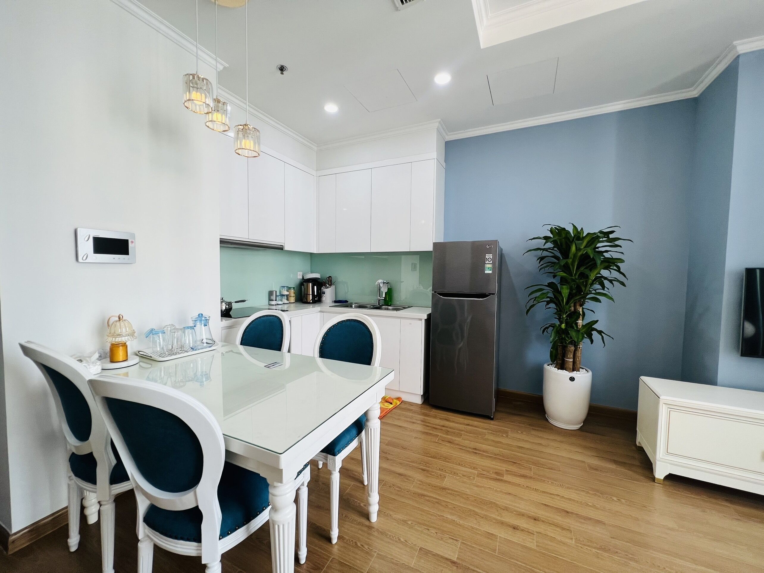 A modern kitchen with white cabinets, a stainless steel refrigerator, a dining table with blue chairs, and a potted plant.