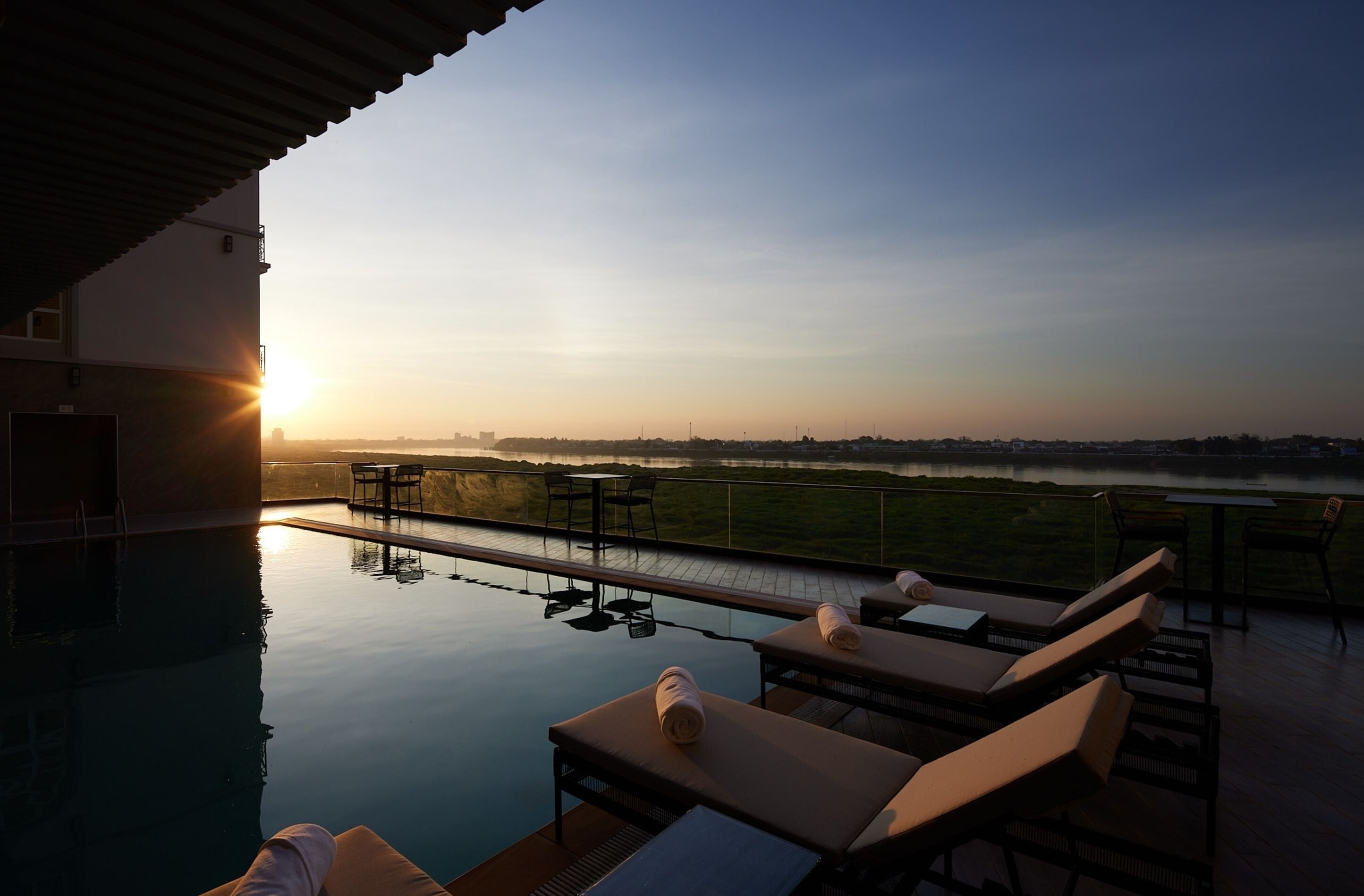 A rooftop infinity pool with lounge chairs and tables, overlooking a body of water and a distant city skyline during sunset.