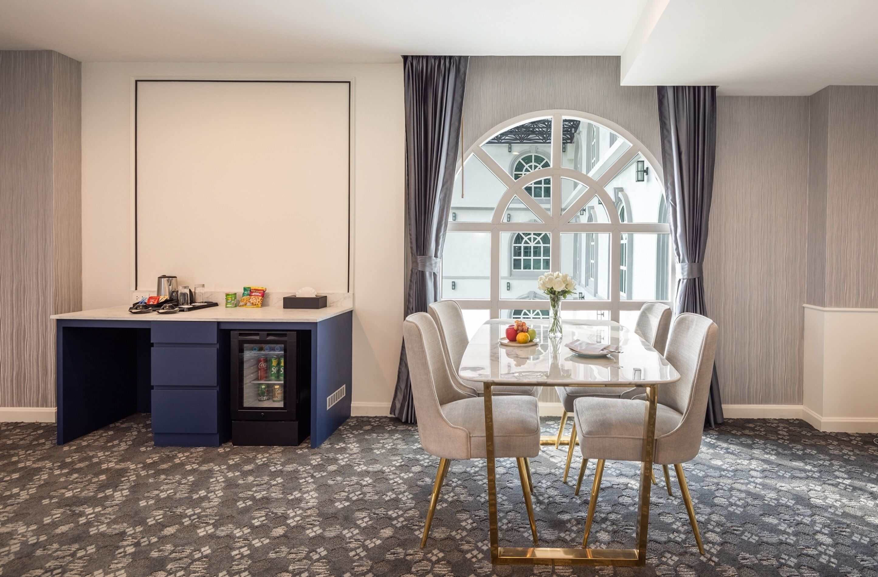 A hotel room with a dining table, chairs, a minibar, and a large window with curtains.