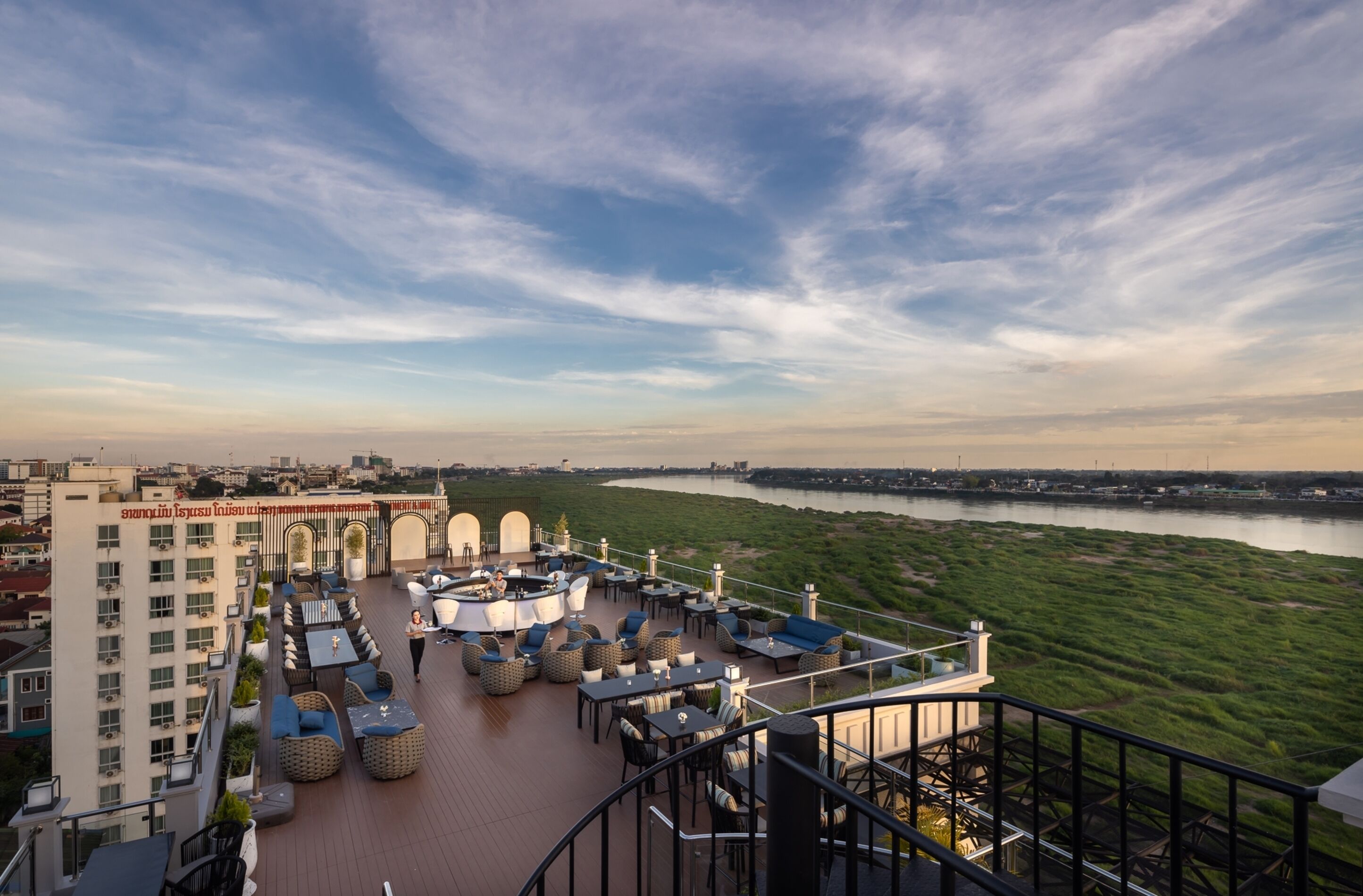 A rooftop terrace with outdoor seating, a view of a river, and a building with a sign in a foreign language.