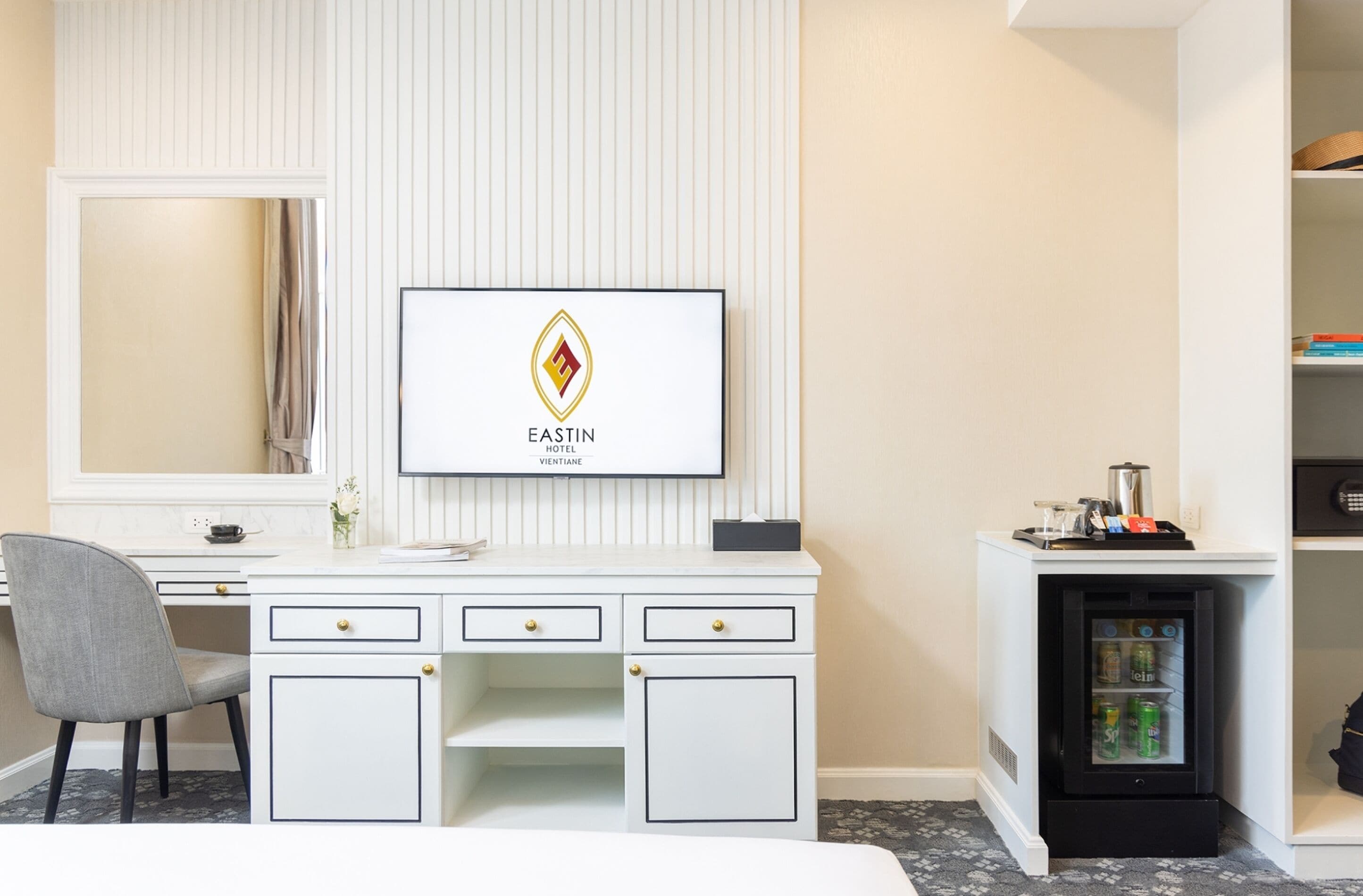 Ein Hotelzimmer mit weißem Schreibtisch, einem Sessel, einem Fernseher, der das Logo des Eastin Hotels zeigt, und einer Minibar.