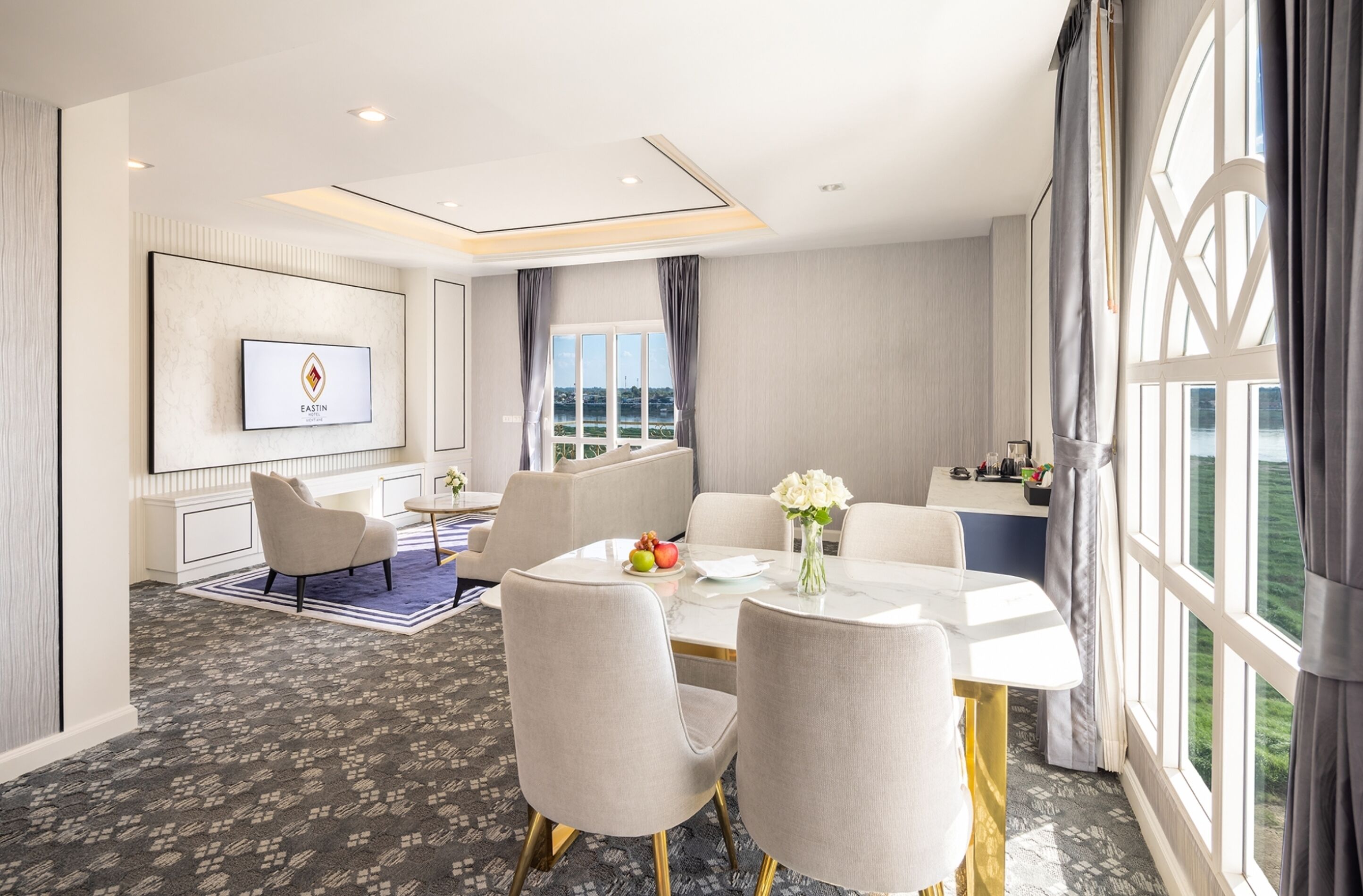 A hotel room with a dining area, a TV mounted on the wall, and a large window with curtains.