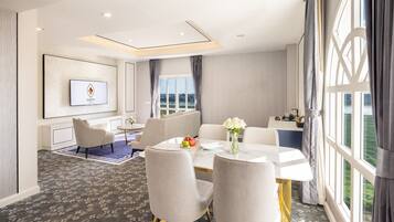 A hotel room with a dining area, a TV mounted on the wall, and a large window with curtains.