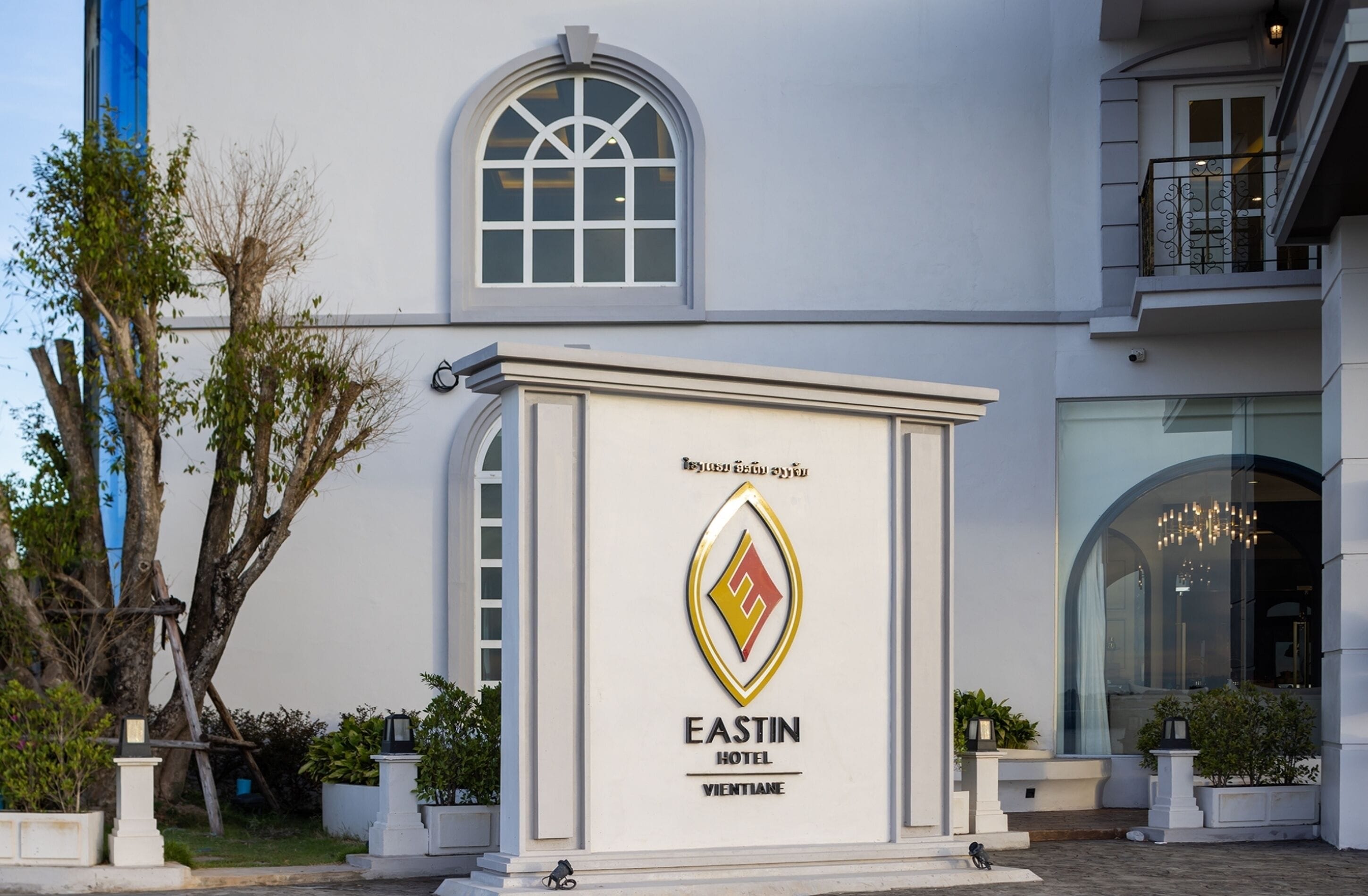 Die Fassade des EASTIN HOTEL VIENTIANE mit einem auffälligen Logo, einem großen Bogenfenster und einem Balkon mit Geländer.