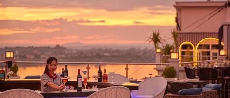 Zona de bar con sillas de mimbre y vista al atardecer sobre un cuerpo de agua.