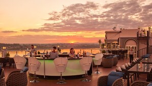 A rooftop bar with wicker chairs and a bar counter, overlooking a sunset over a body of water.